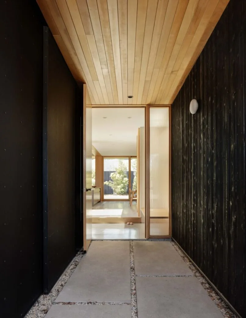 Entry corridor with wood ceiling and charred wood siding opens to a sunlit interior featuring large windows toward a courtyard.