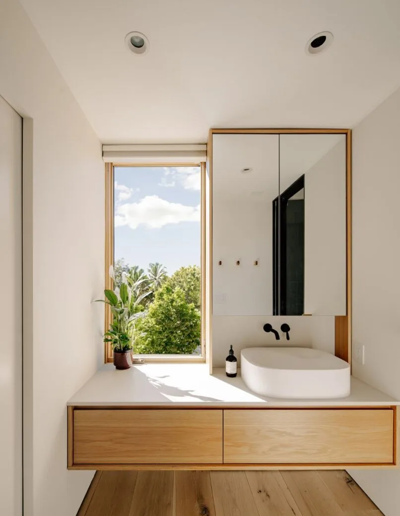 Bathroom vanity with wood-paneled drawer, wall-mounted faucet, and tall window overlooking greenery in Cupertino Courtyard House.