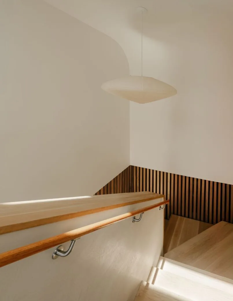 Staircase in Cupertino Courtyard House features vertical wood slat wall treatment and light wood handrail under a beige pendant light.