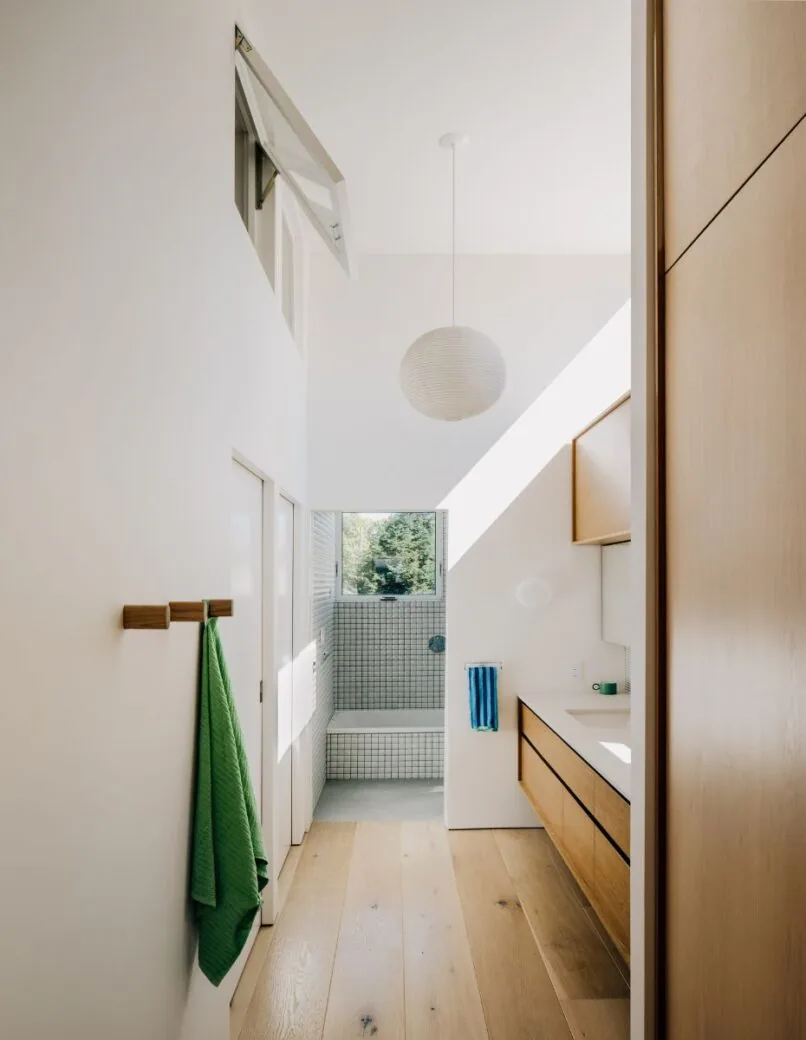 Narrow bathroom features wood flooring, floating vanity with wood drawers, grid-tiled shower area, and a round pendant light.