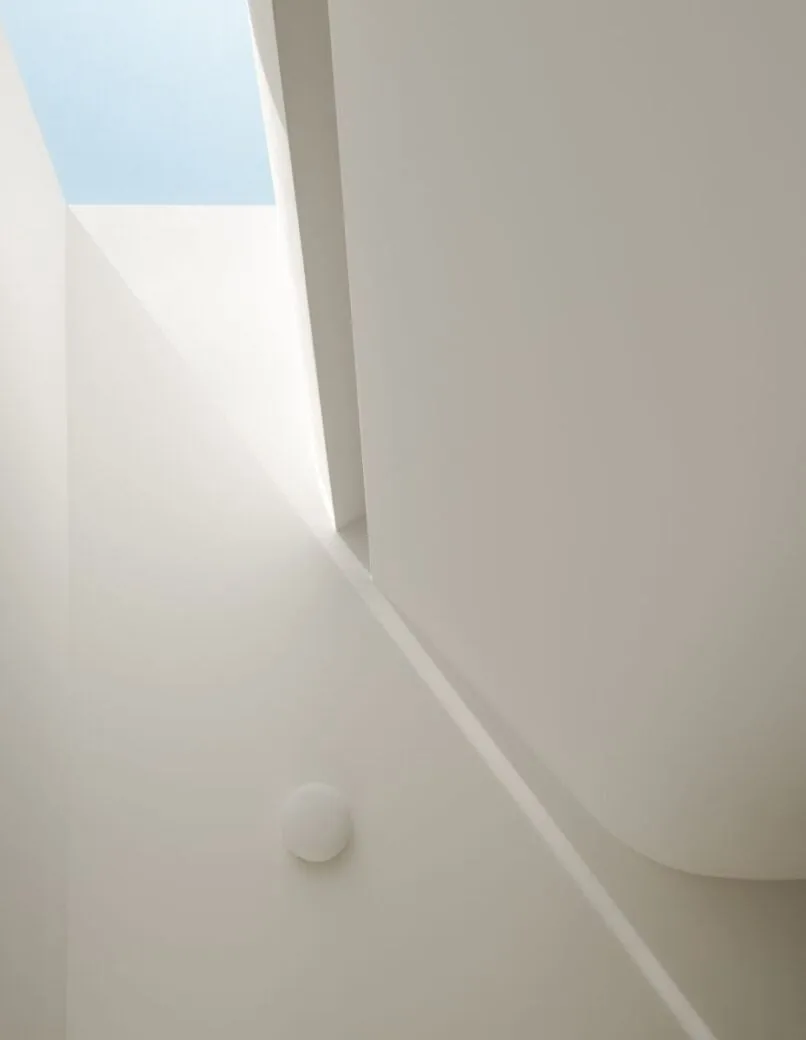 White stairwell with a round wall light and a rectangular skylight showing clear blue sky.