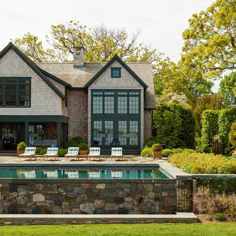Shingle-style house features large green-framed French doors opening to a pool patio with lounge seating and surrounding landscaping.