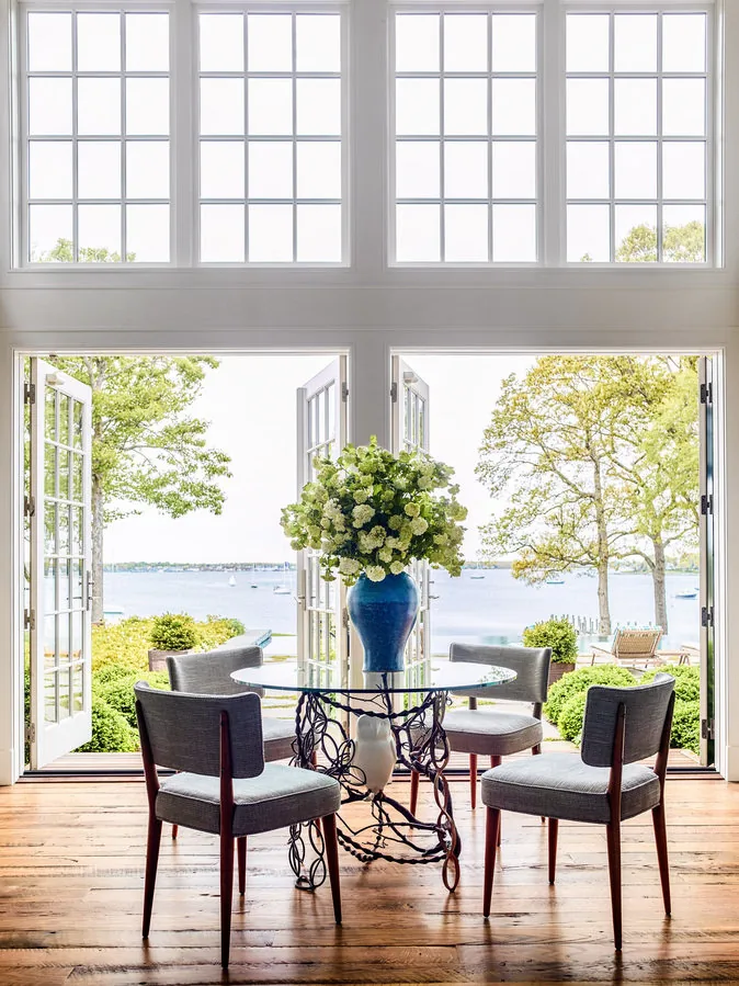 Dining area with a glass table and four chairs opening to a view of a waterfront through French doors with multiple panes.