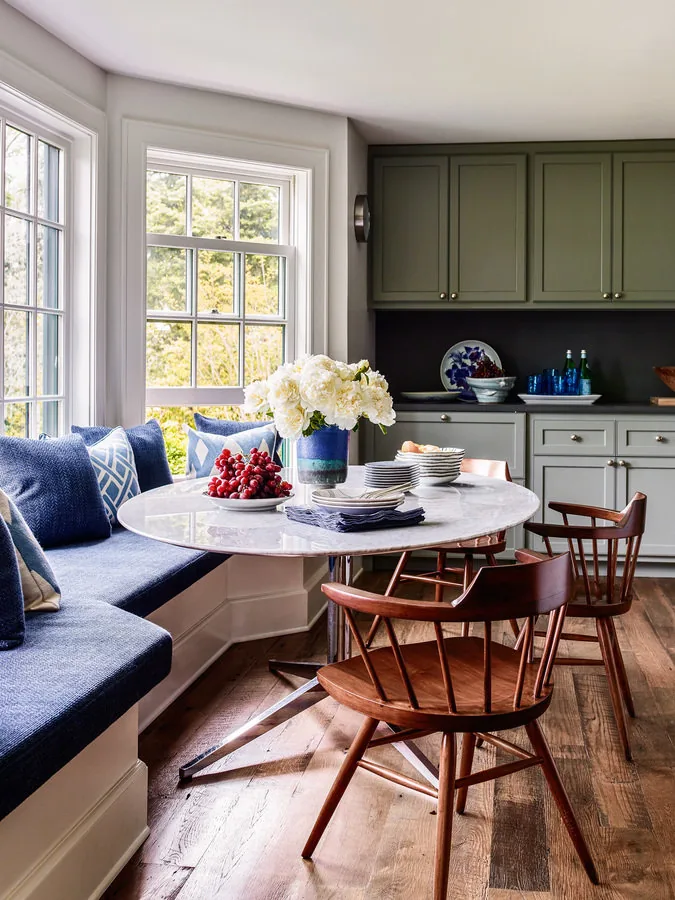 Kitchen dining nook with green cabinets, blue cushioned bench, and wooden chairs around a round marble table.