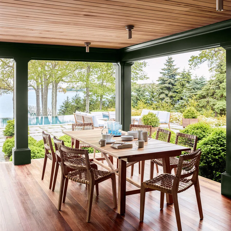 Outdoor dining space features wooden furniture under a covered patio with green beams and a waterfront backdrop.