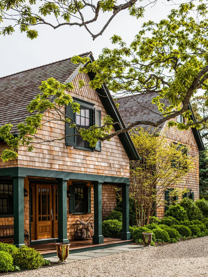 Exterior view of a traditional wooden house with cedar shingle siding and dark green trim, surrounded by leafy green trees.