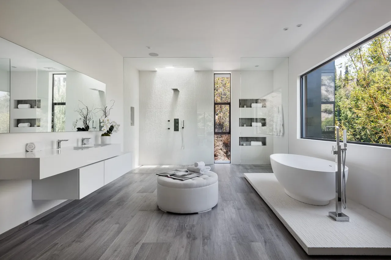 This modern bathroom features a freestanding tub on a platform, a walk-in shower with tall narrow window, and a floating double vanity with mirror.