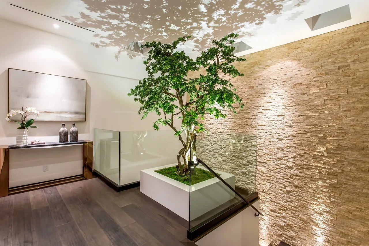 An interior stair landing featuring a tree planted in a white planter and illuminated textured stone wall.
