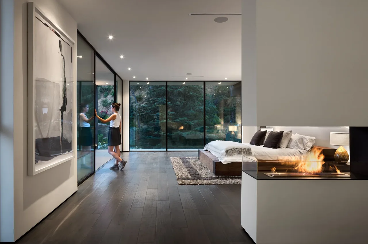 Bedroom featuring large glass windows, a modern fireplace, and dark wood flooring in the Rising Glen project by MSP Design Development.