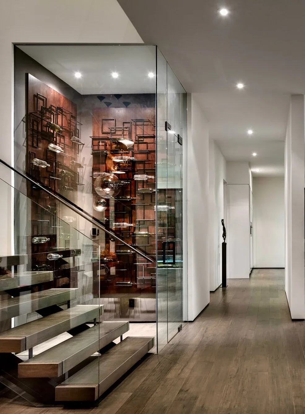 A modern hallway features floating wood stairs and a glass-enclosed, wall-mounted wine rack.