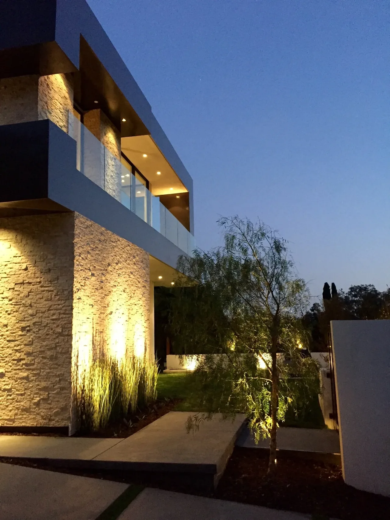 Modern two-story house featuring stone walls, glass balcony, and exterior lighting at dusk.