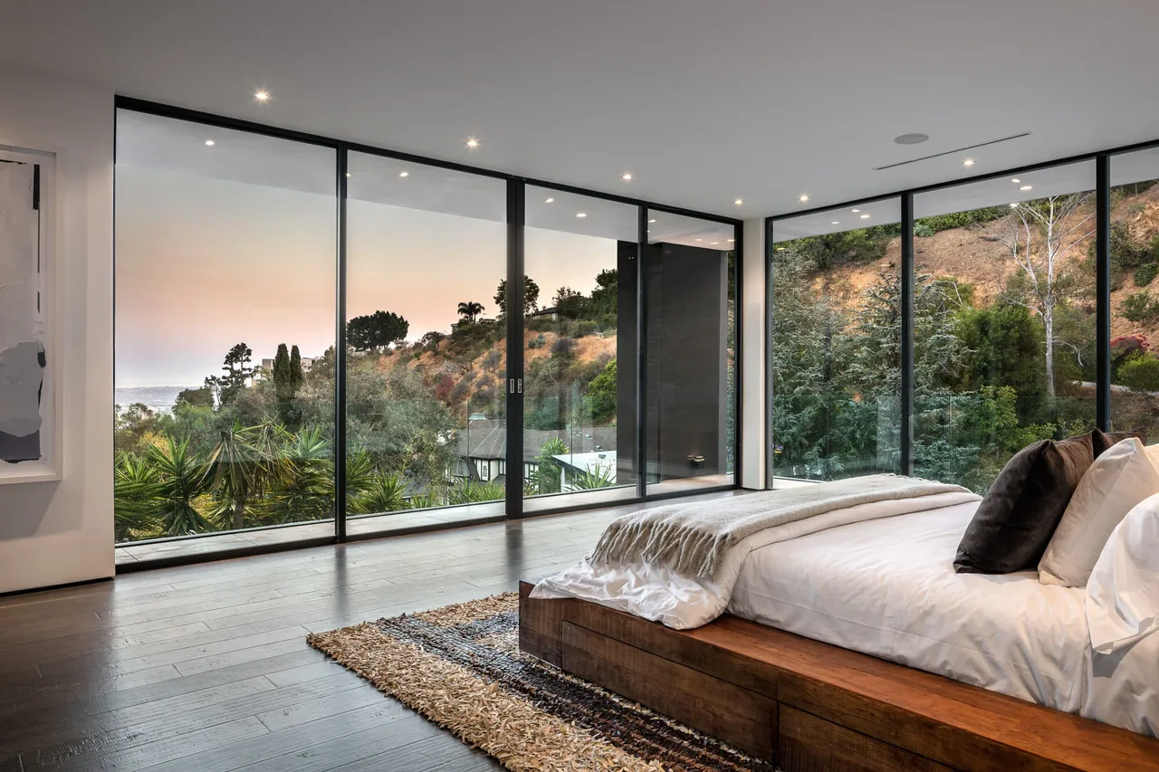 Bedroom features floor-to-ceiling black-framed glass windows with hillside views and recessed ceiling lighting.