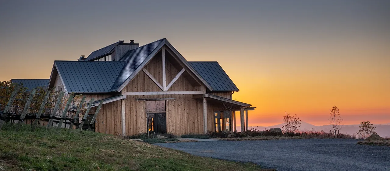 The Crimson Lane Vineyard building features wood siding and a standing seam metal roof with A-frame trusses visible at sunset.