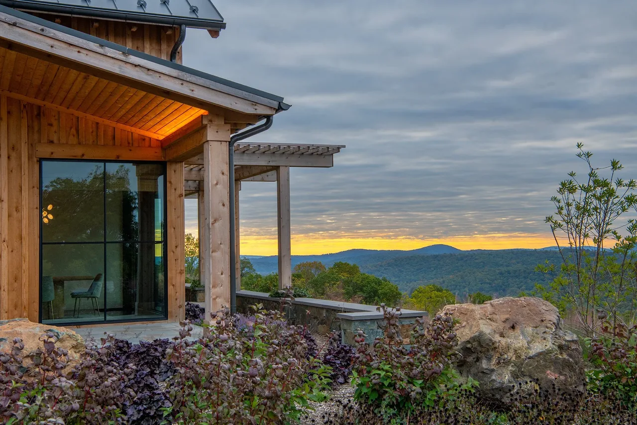 Wood-clad home corner with black-framed floor-to-ceiling glass window overlooks landscape at sunset.
