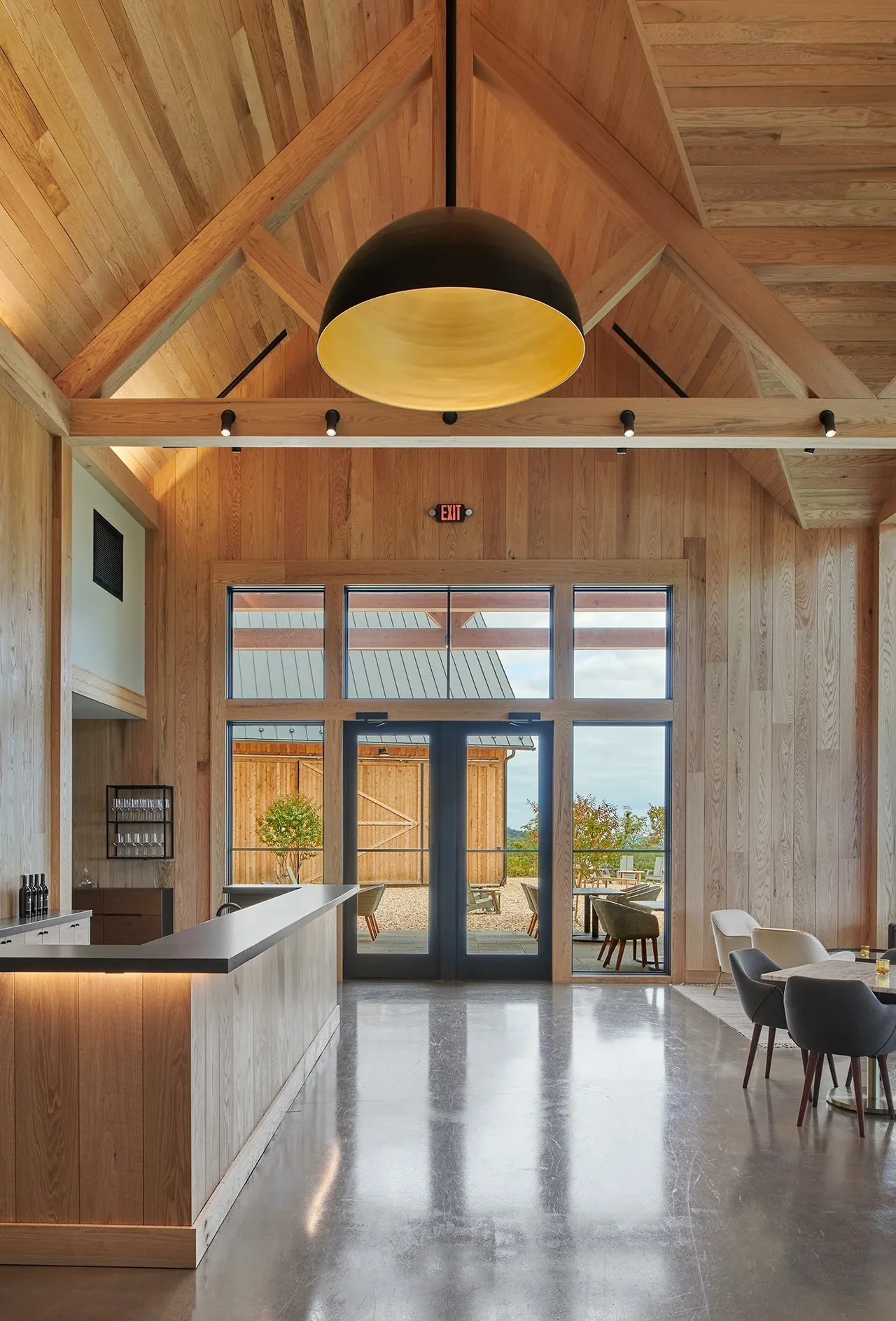 Timber-paneled vaulted ceiling living space with a large black pendant light and double glass doors opening to outdoor seating.