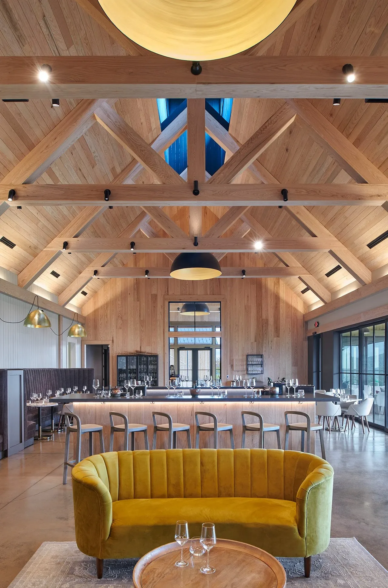 The vaulted ceiling space features wood paneling, a yellow curved sofa, and a bar counter lined with chairs, combining rustic and modern elements.