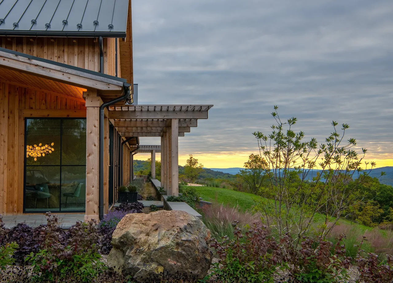 Timber-framed house exterior with a metal roof and pergola overlooking a natural stone and gravel landscape at sunset.