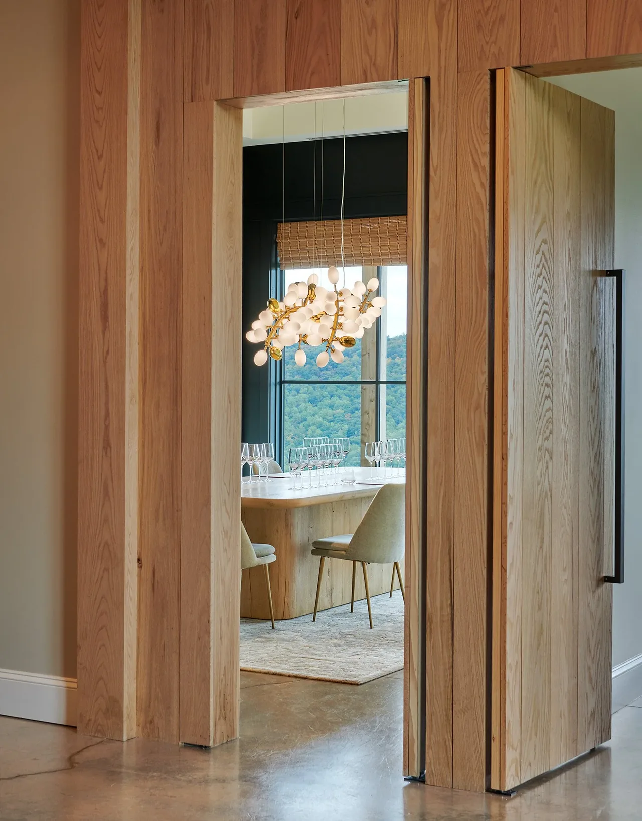 Dining room seen through wood-paneled pivot doors featuring upholstered chairs and a sculptural pendant light.