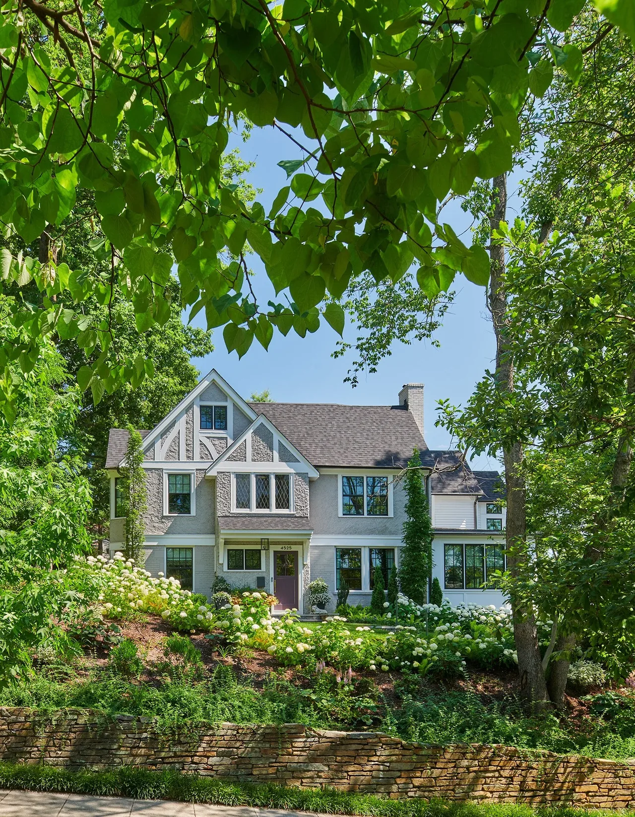 Tudor-style home with textured exterior and multi-pane windows set behind mature trees and landscaped flower beds.