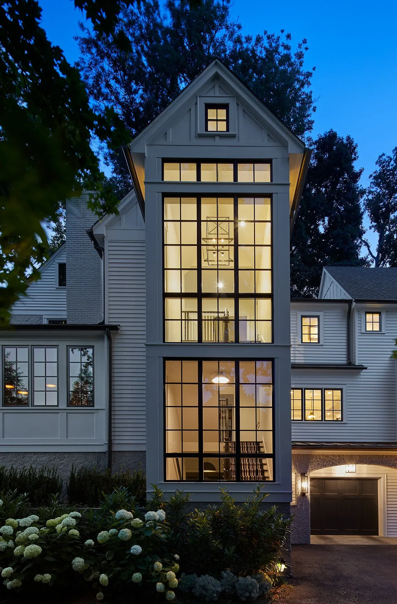 Traditional styled house exterior with gable roof and large grid windows illuminating an interior stairway at dusk.