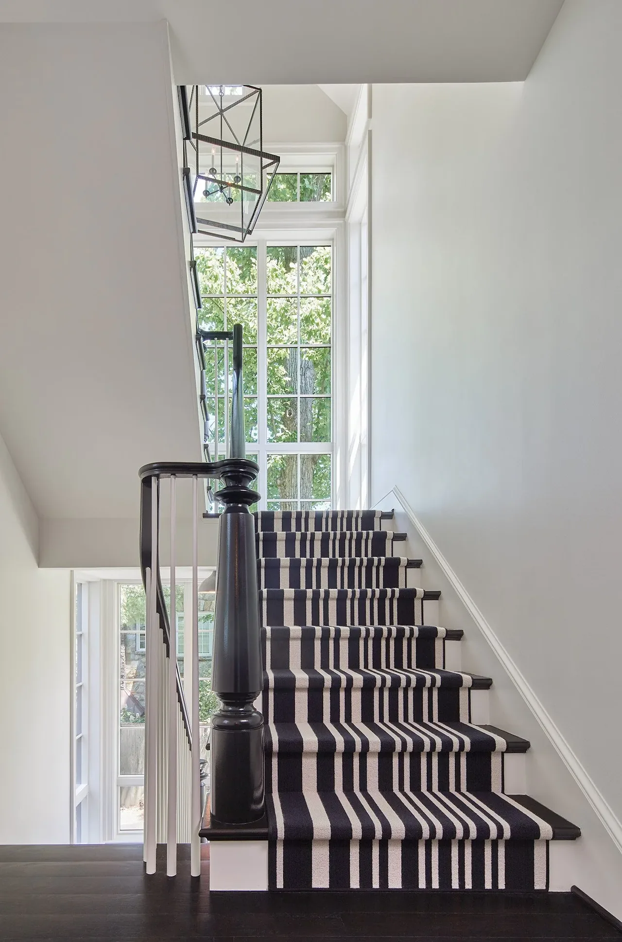 Stairway with a striped carpet runner and black wood handrail next to large multi-pane leaded windows.