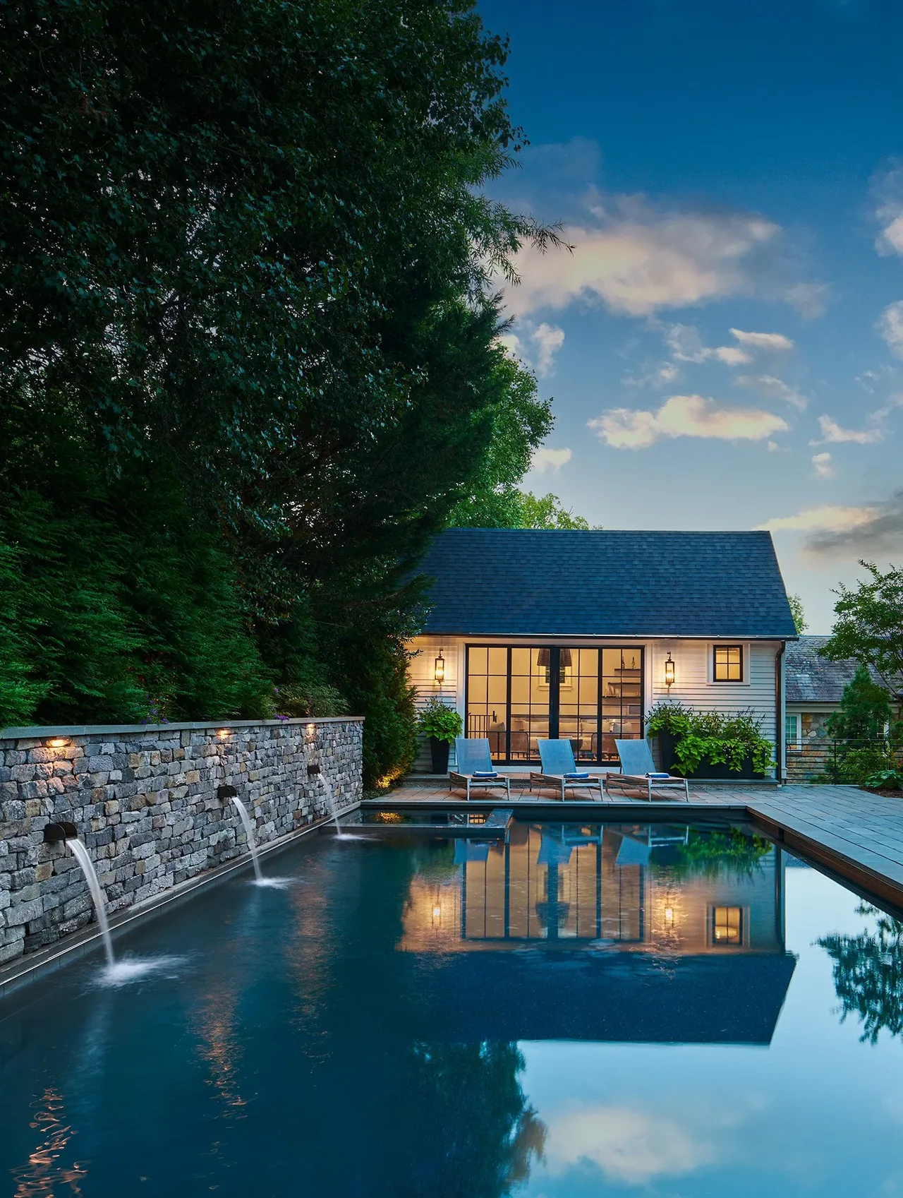 Stone wall with water spouts beside a rectangular pool reflects a white pool house with black-framed glass doors at dusk.