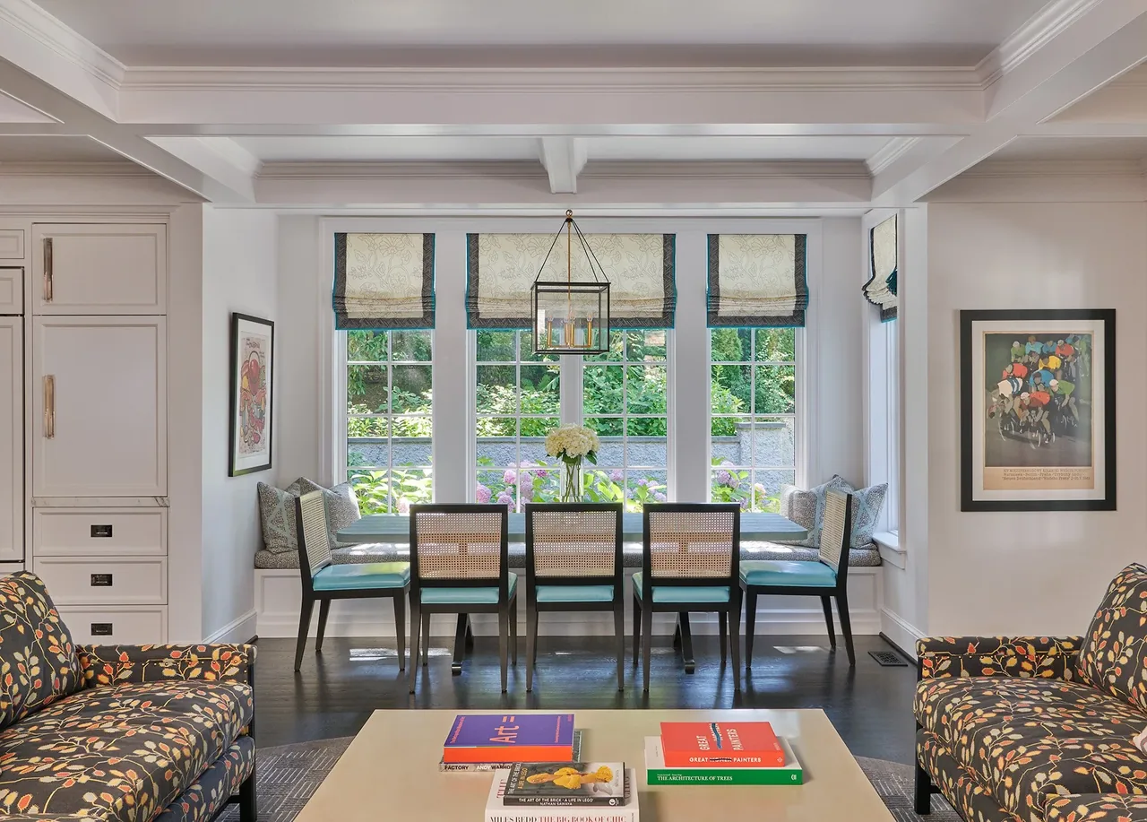 Dining space features a rectangular table with six cane-back chairs and built-in bench under multi-pane windows with roman shades.