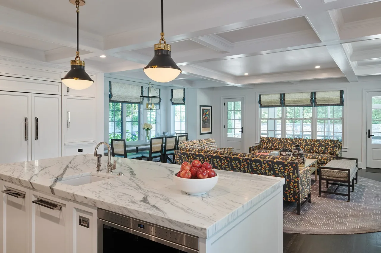 Interior view of a kitchen with marble island and adjacent living room with floral sofas and large windows.