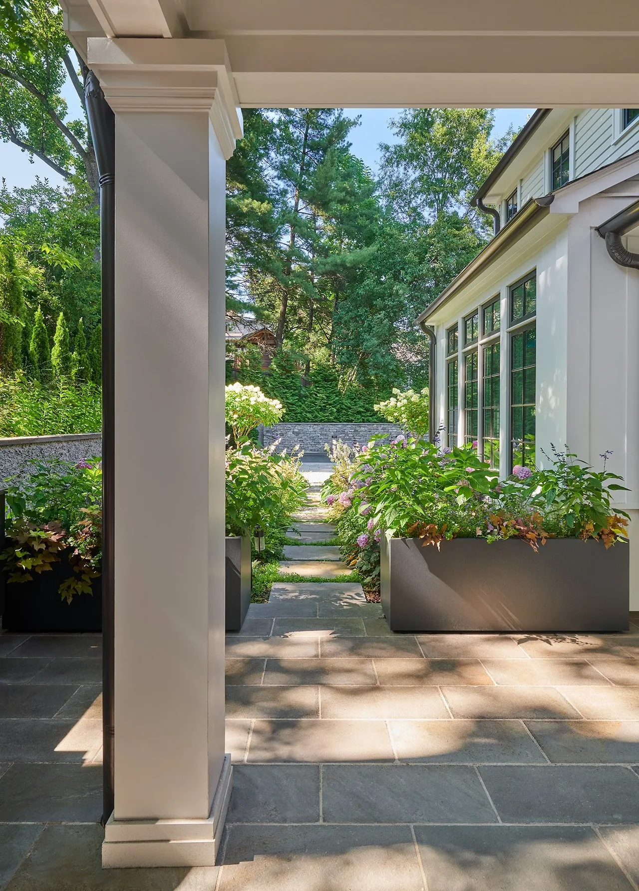 Covered patio with stone flooring, large planters, and landscaped garden visible beyond a multi-pane windowed house exterior.