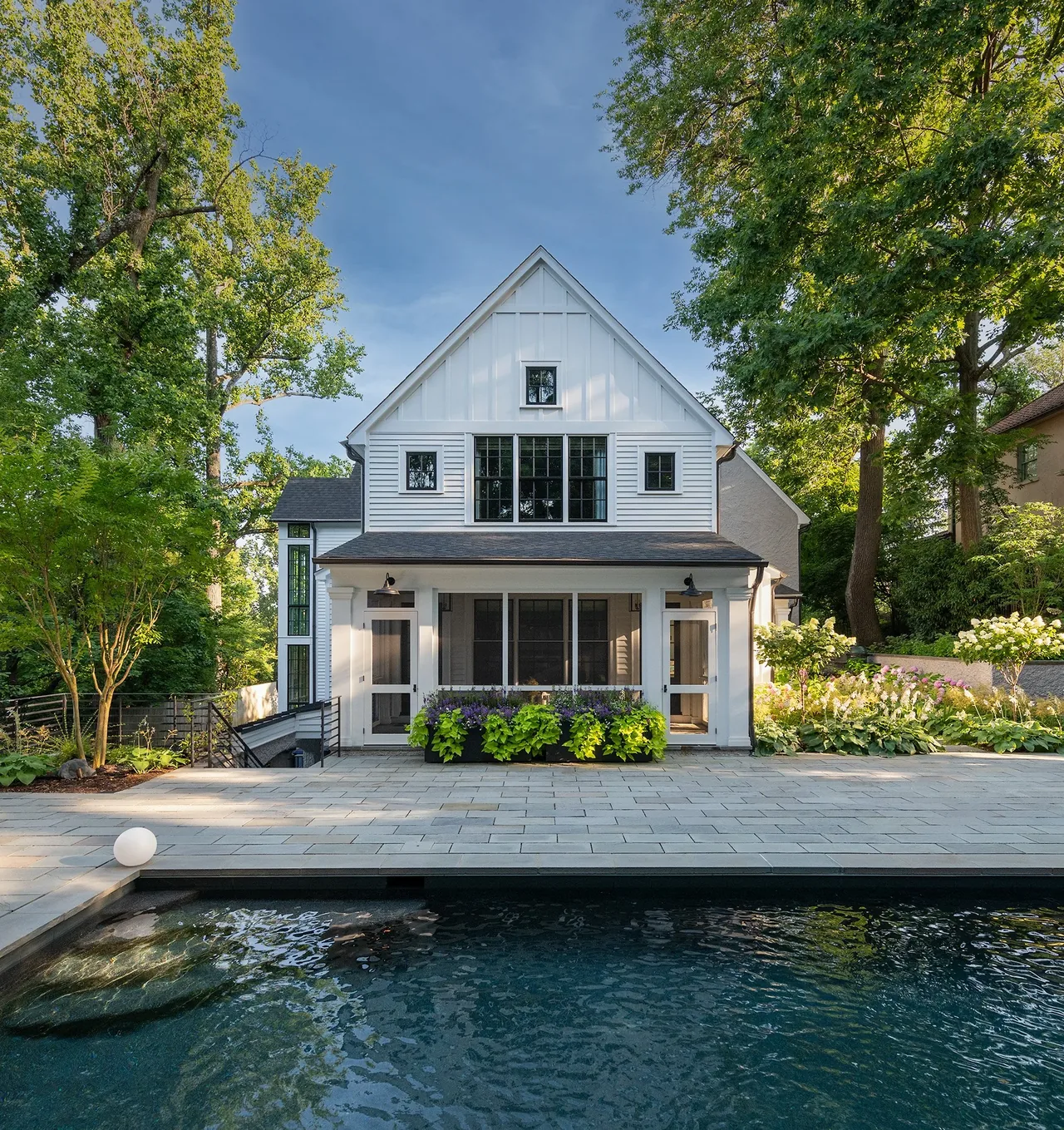 Back view of a modern farmhouse with symmetrical multi-pane windows and a gable roof adjacent to a pool area with paved stone flooring.