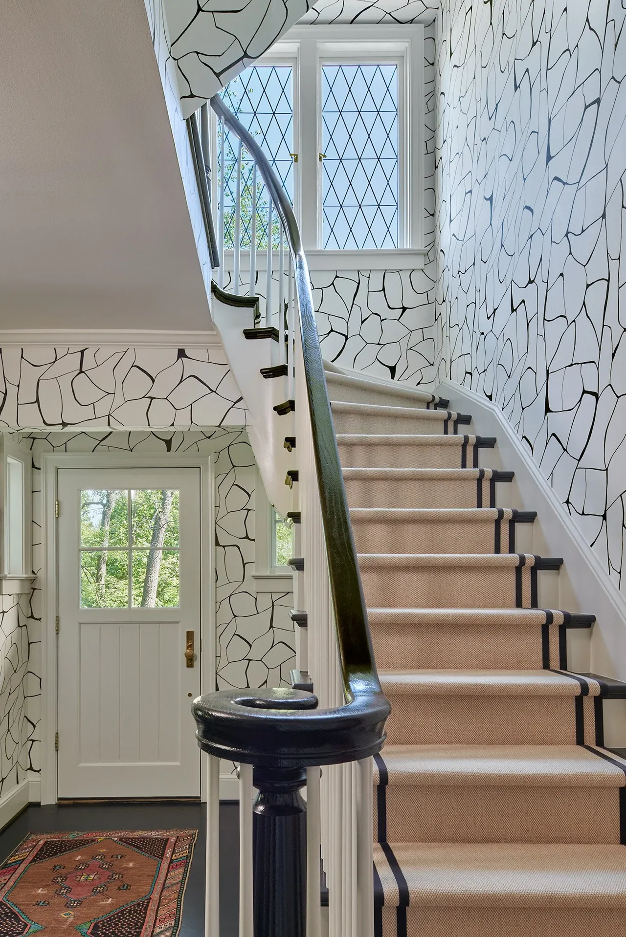 Staircase features a beige carpet runner with black stripes and leaded diamond pane windows above the door.