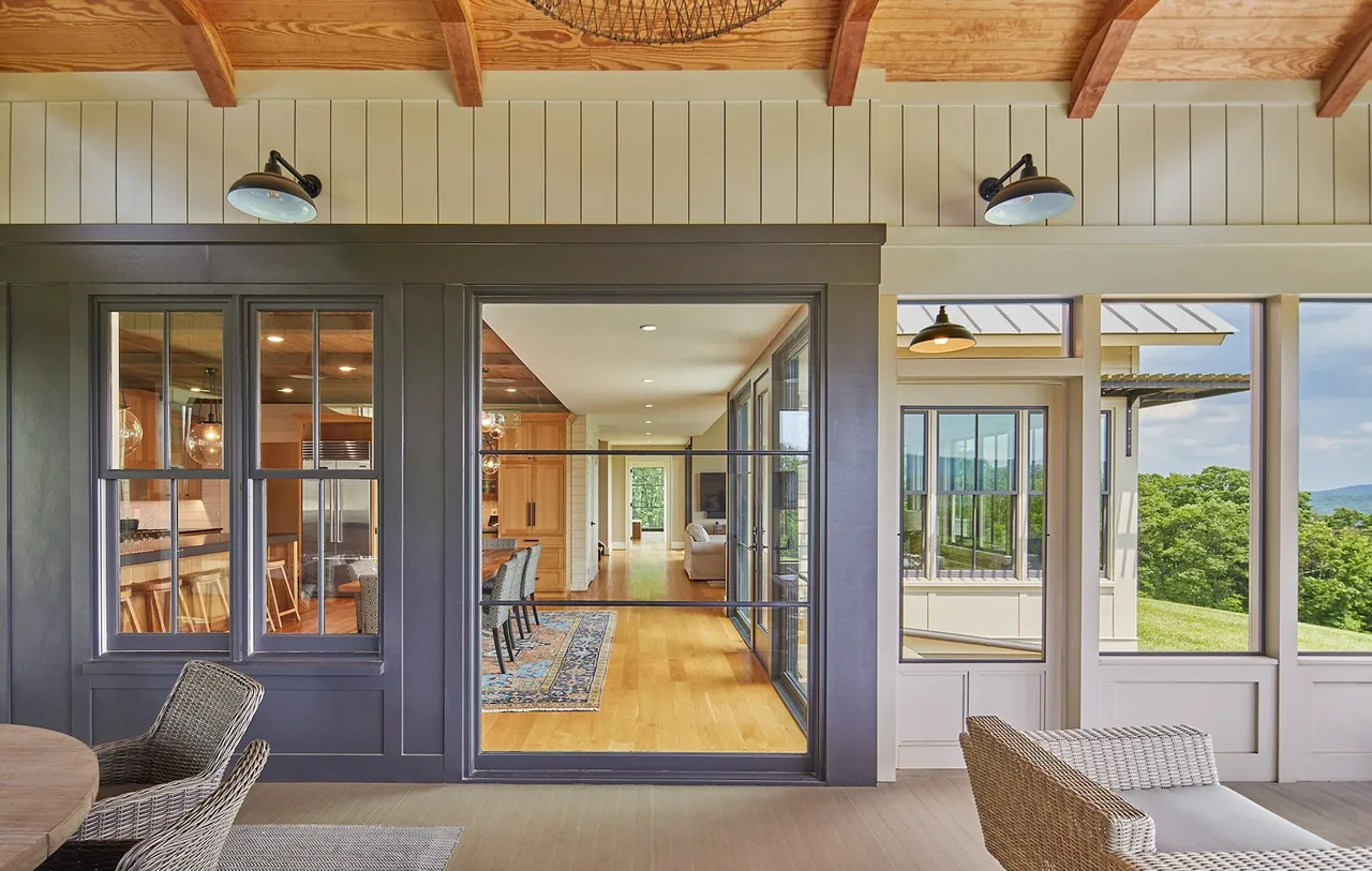 Covered porch with wicker furniture overlooks an interior with hardwood floors and dark gray framed windows and doors.