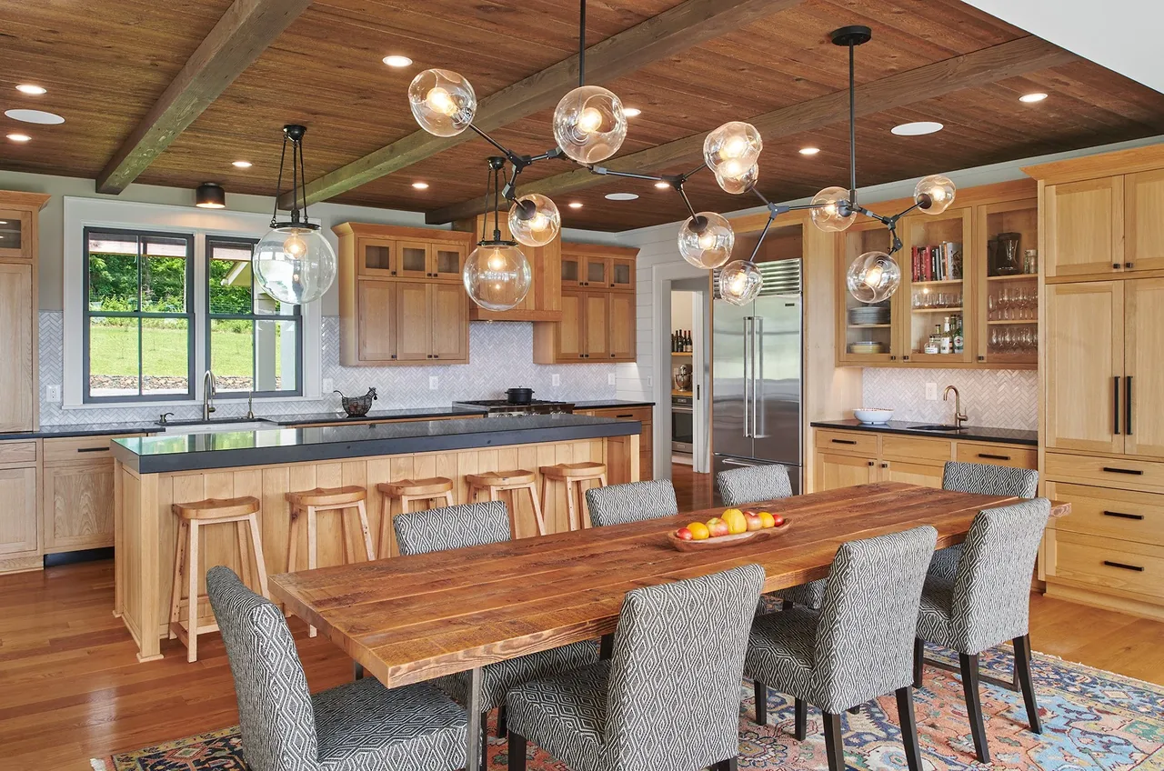 Kitchen and dining space feature wood cabinetry, a long island, and a large wooden table with patterned chairs under globe pendant lights.