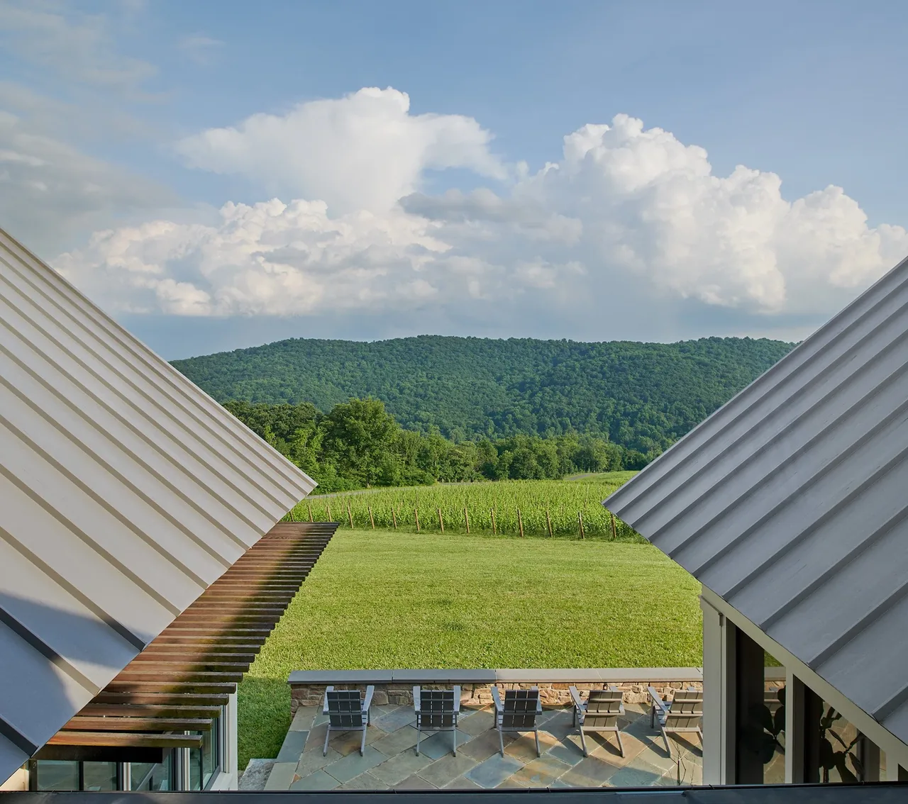 Stone patio with seating between two buildings with standing seam metal roofs overlooks a lush green field and hills.