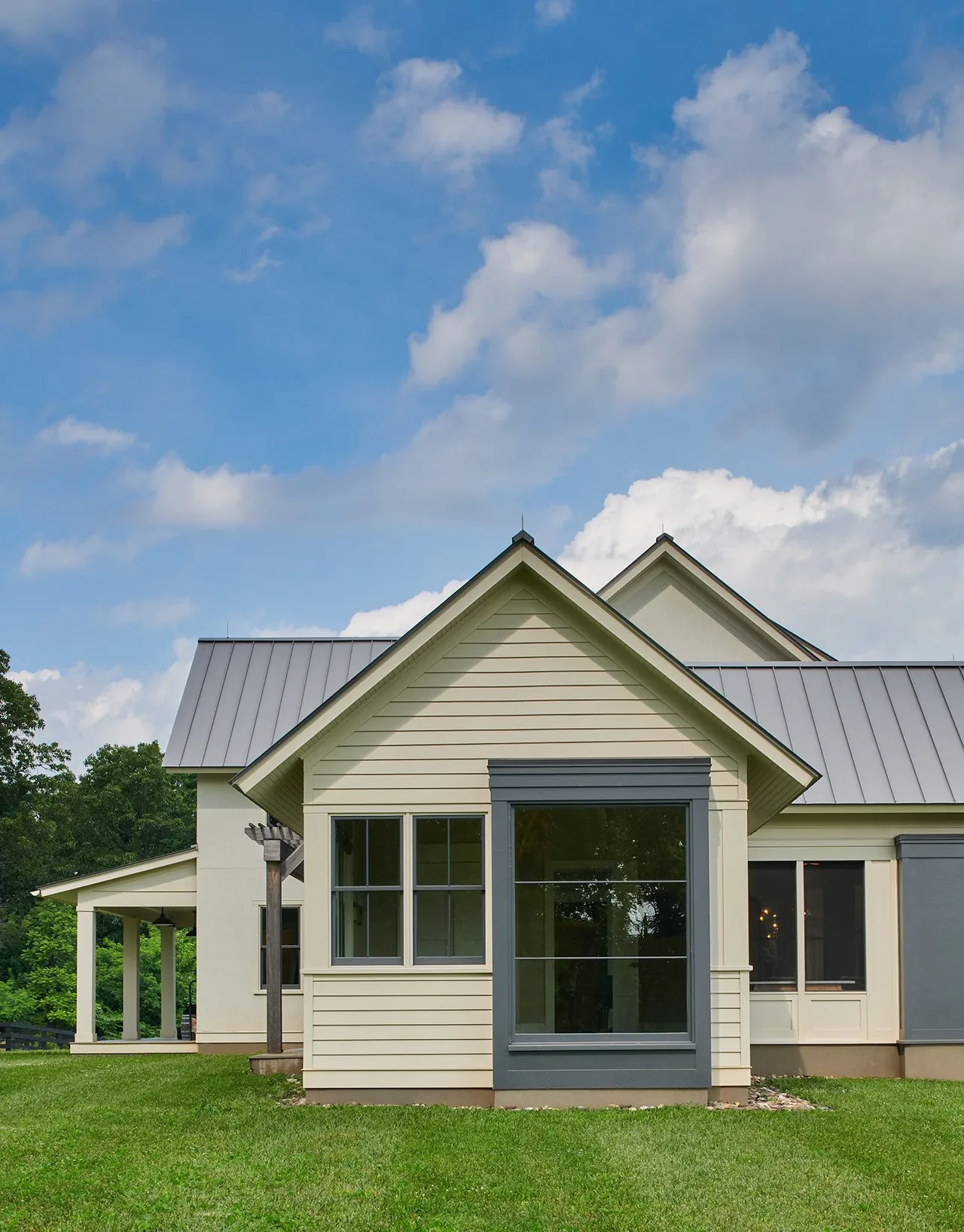 The Crimson Lane Residence features light beige siding, large gray-framed picture windows, and a standing seam metal roof.