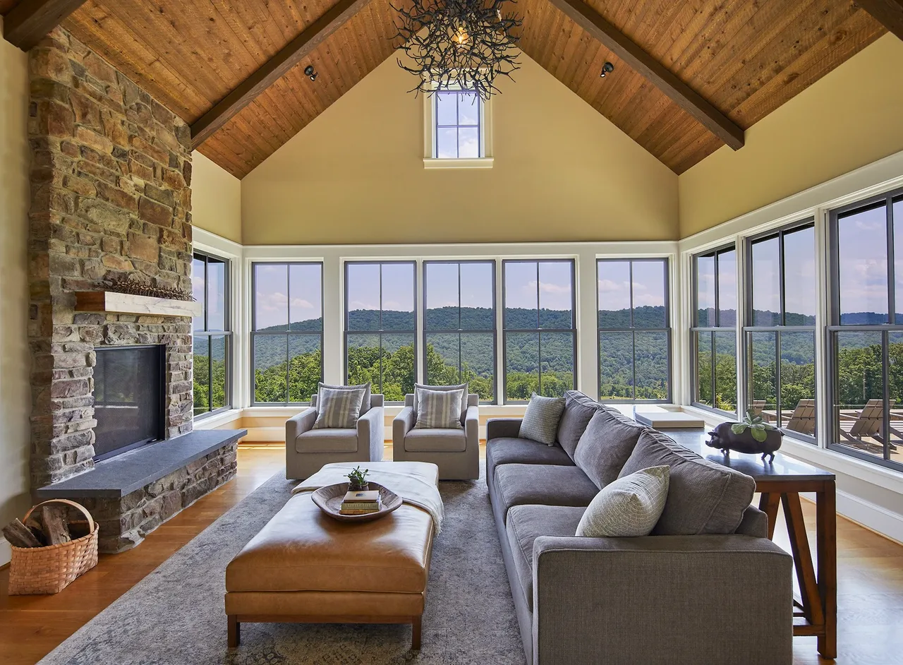 Living room with vaulted wood ceiling and stone fireplace overlooks scenic hills through tall windows.
