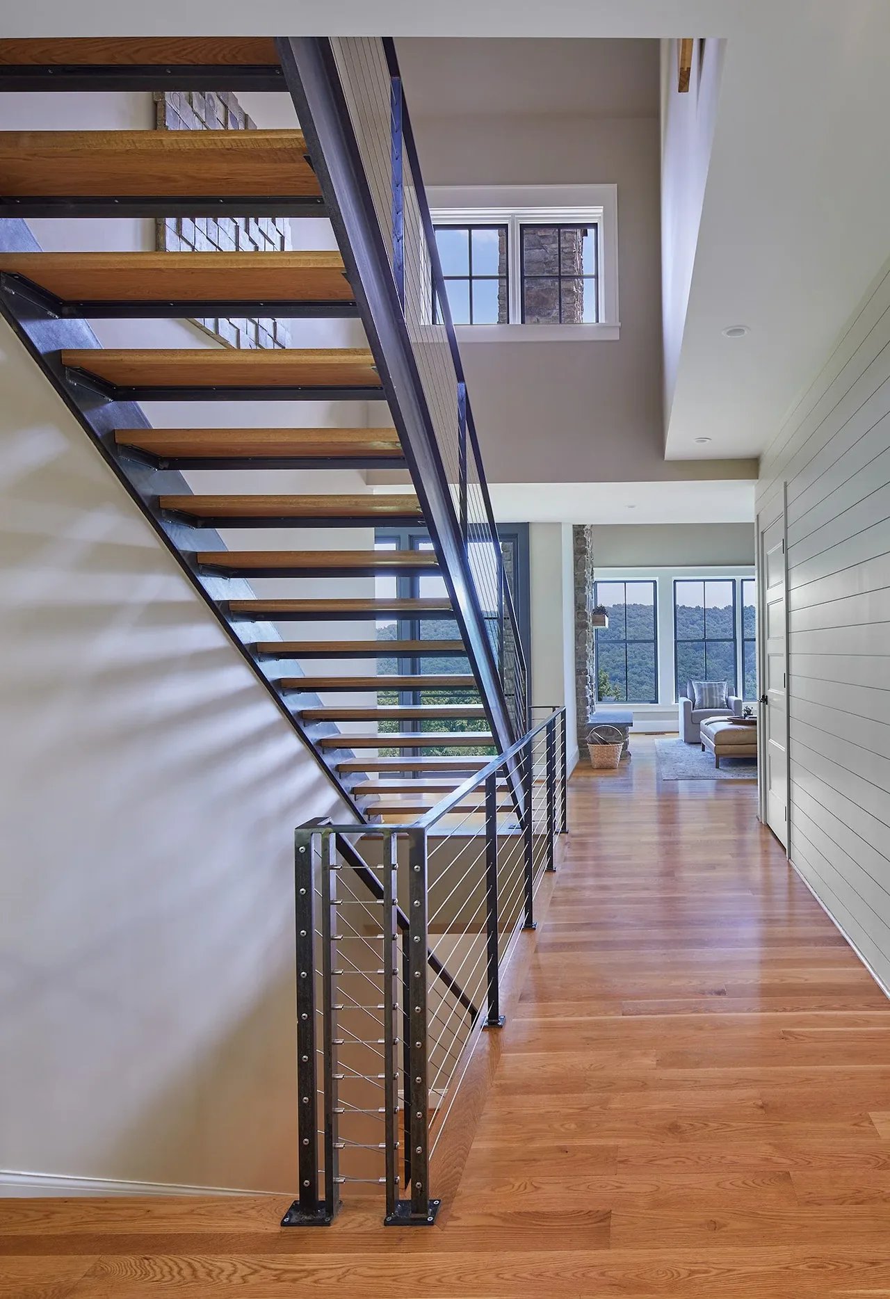 Wood and metal floating stairs with cable railings in a hallway overlooking a living area with large windows.