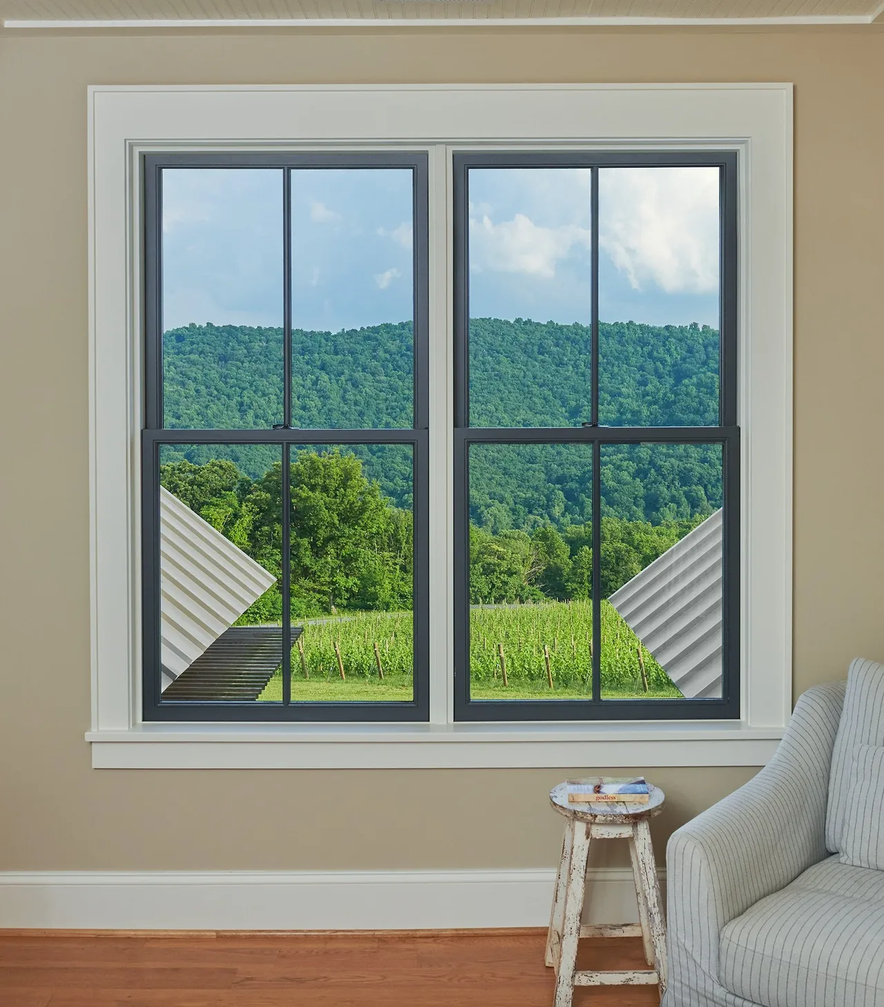 Interior view shows a double black-framed window with a green hillside outside, accompanied by a striped armchair and rustic stool indoors.