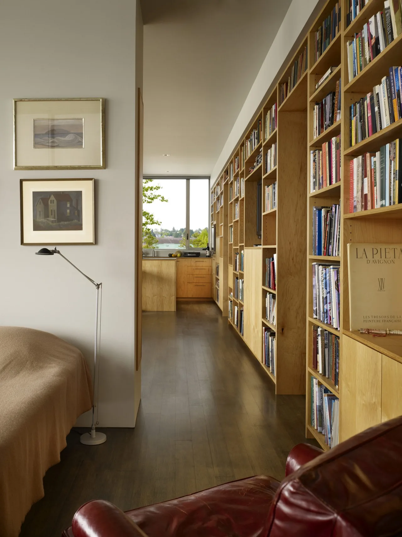 Built-in wooden bookshelves line a hallway that leads to a kitchen with a large window overlooking greenery.