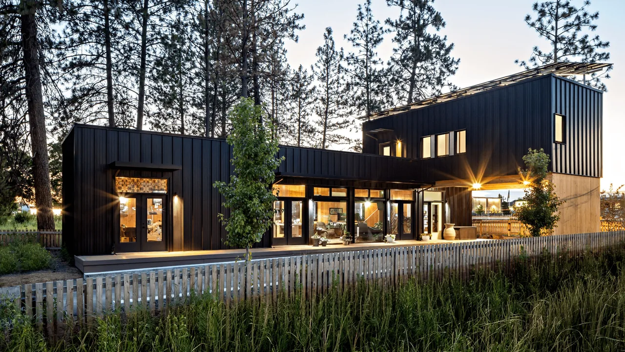 Evening view of a two-story modern house with black standing seam metal siding and large glass doors surrounded by trees and a wooden fence.