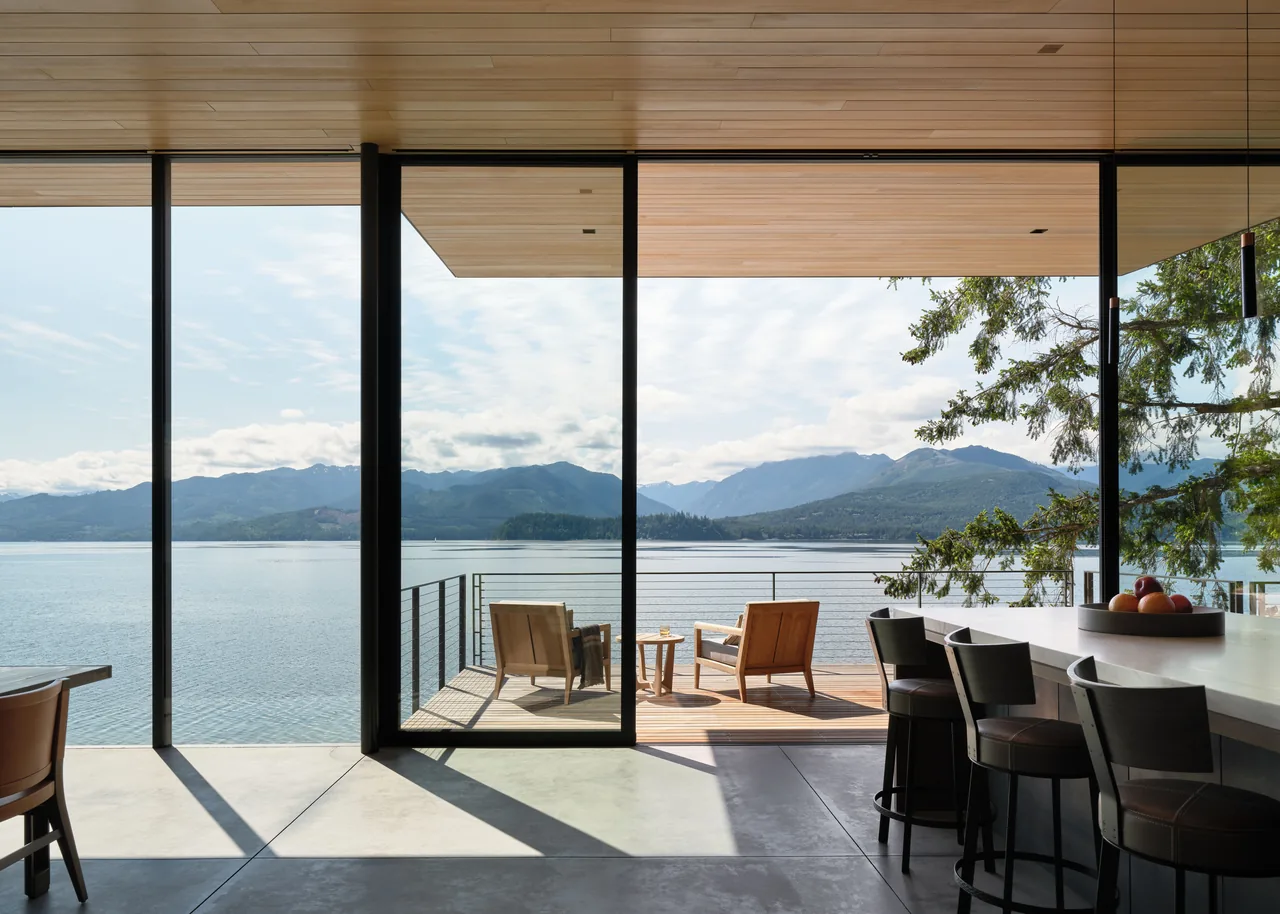 Interior view of a kitchen with floor-to-ceiling glazing overlooking a lake and mountains from an outdoor seating deck.