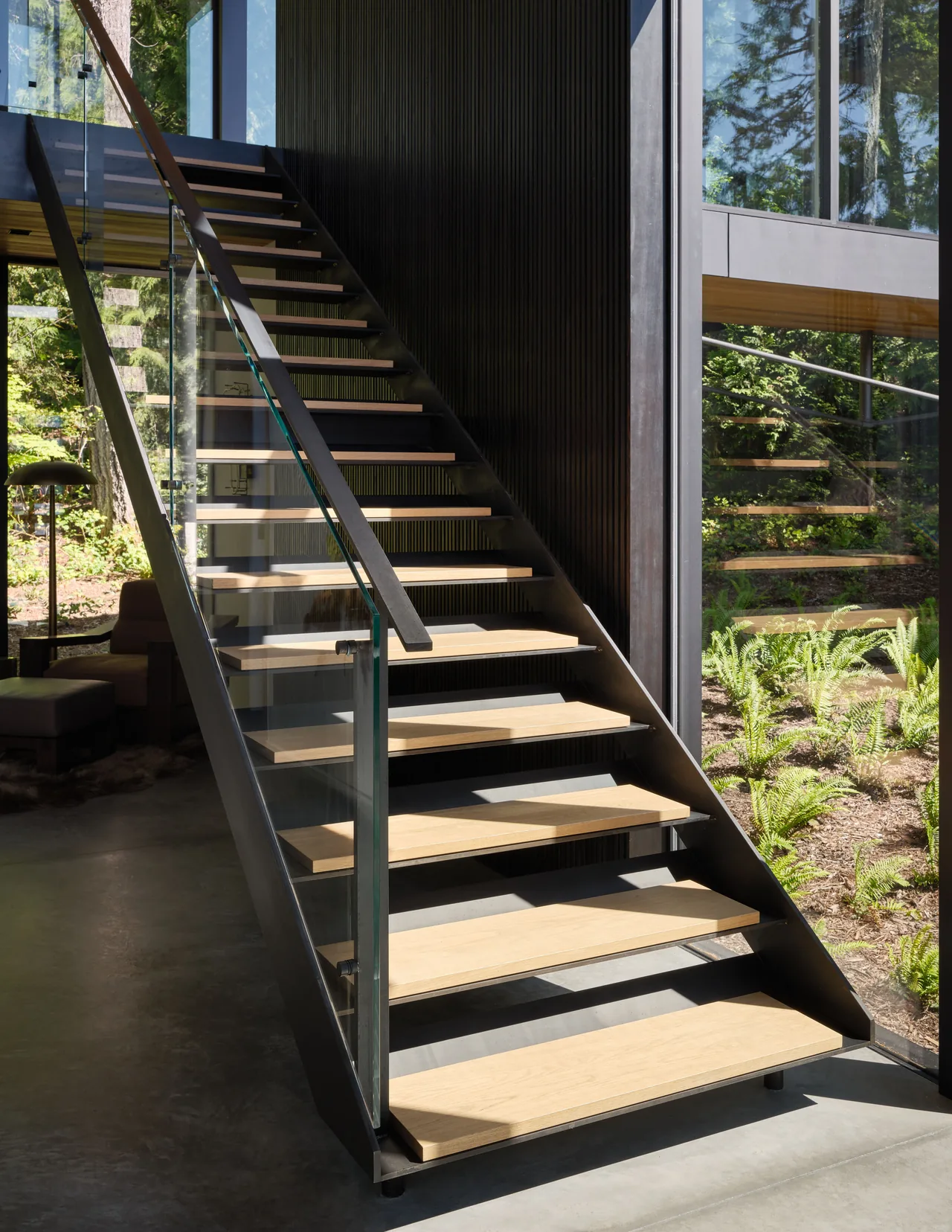 Floating staircase with wooden treads and glass railings in a modern home interior next to large windows.