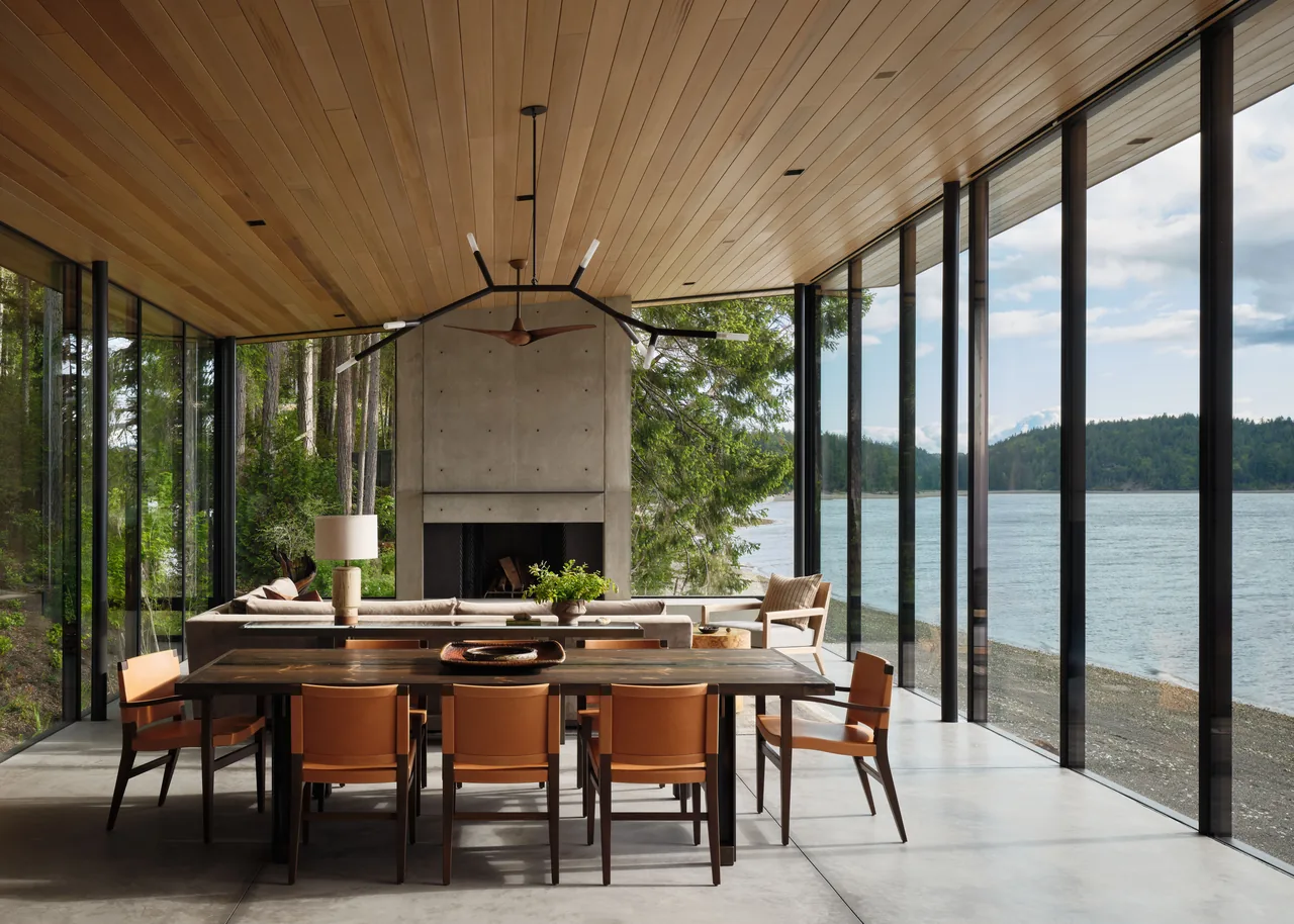 Living and dining room with timber ceiling, concrete fireplace, and floor-to-ceiling glazing overlooking a lake.