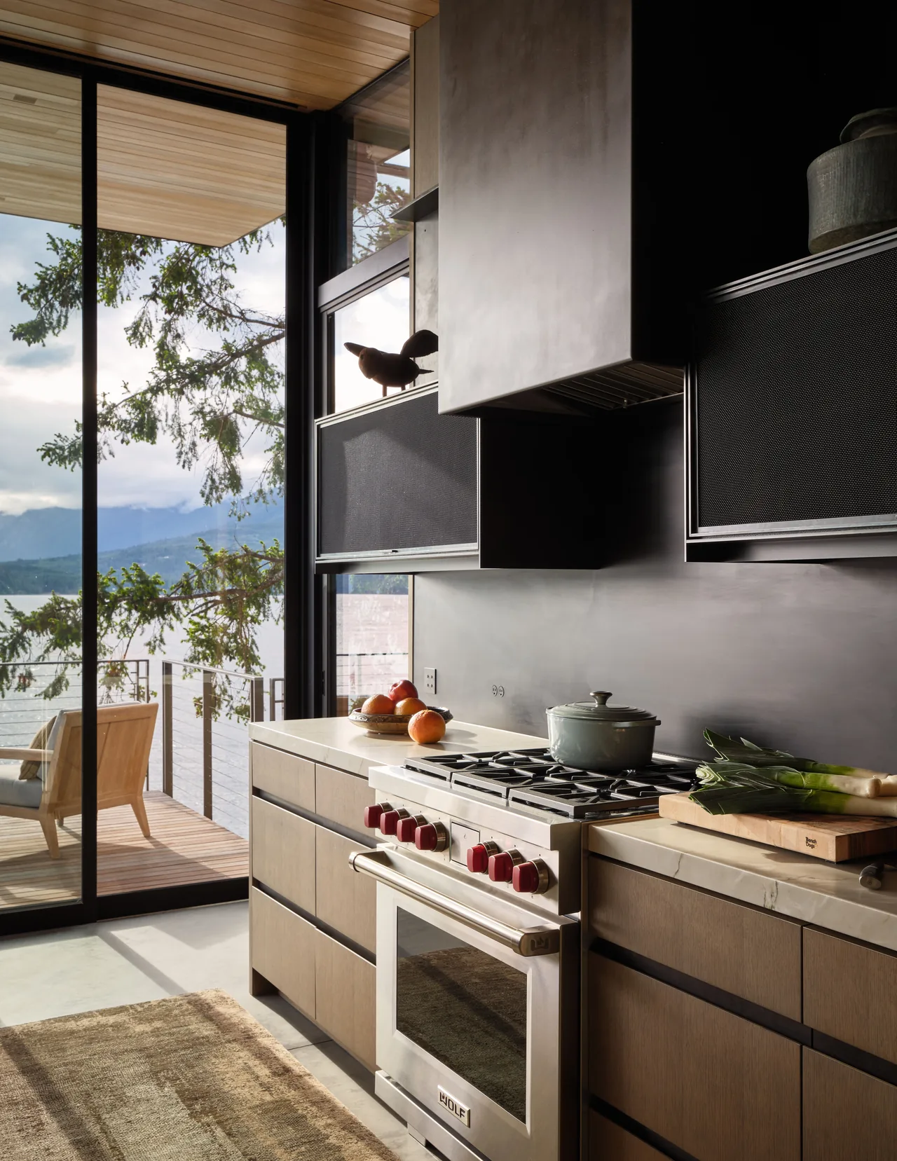 This kitchen features a stainless steel gas range with red knobs, wood cabinetry, and floor-to-ceiling glass doors opening to a deck with forest views.