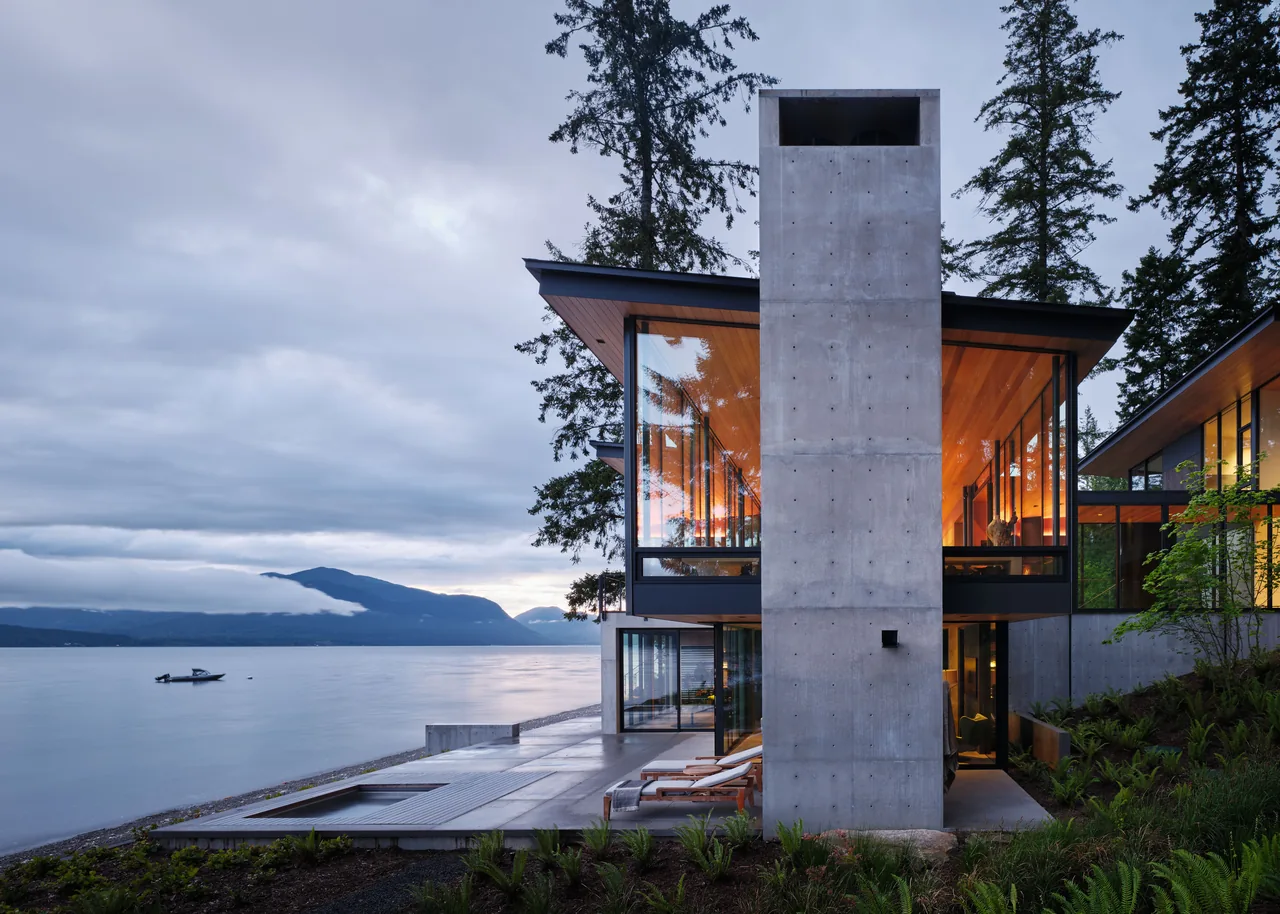 Modern concrete home with floor-to-ceiling glazing and flat roof at lakefront during dusk.