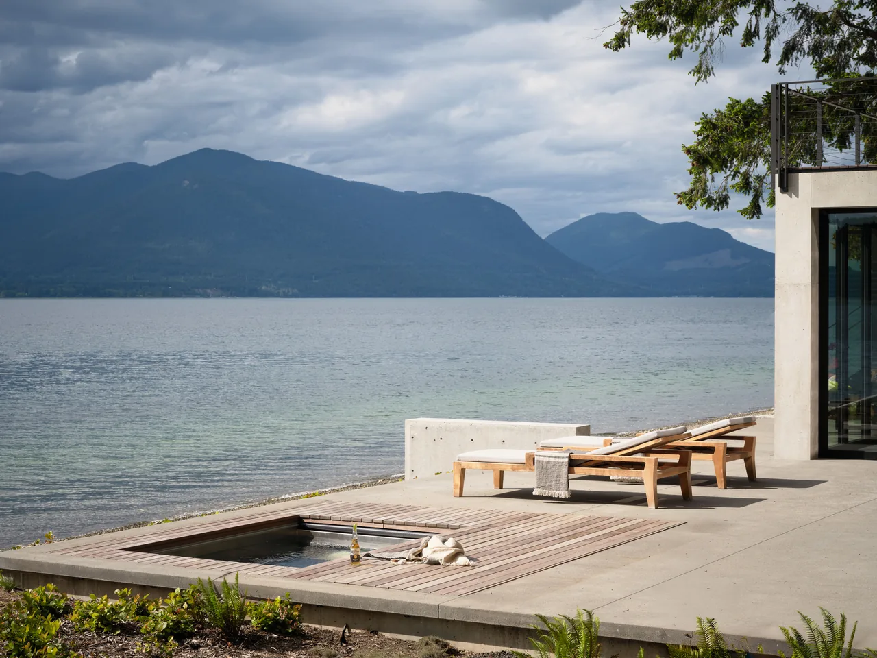 Concrete patio beside a lake features two wooden lounge chairs and a recessed hot tub with mountain views.