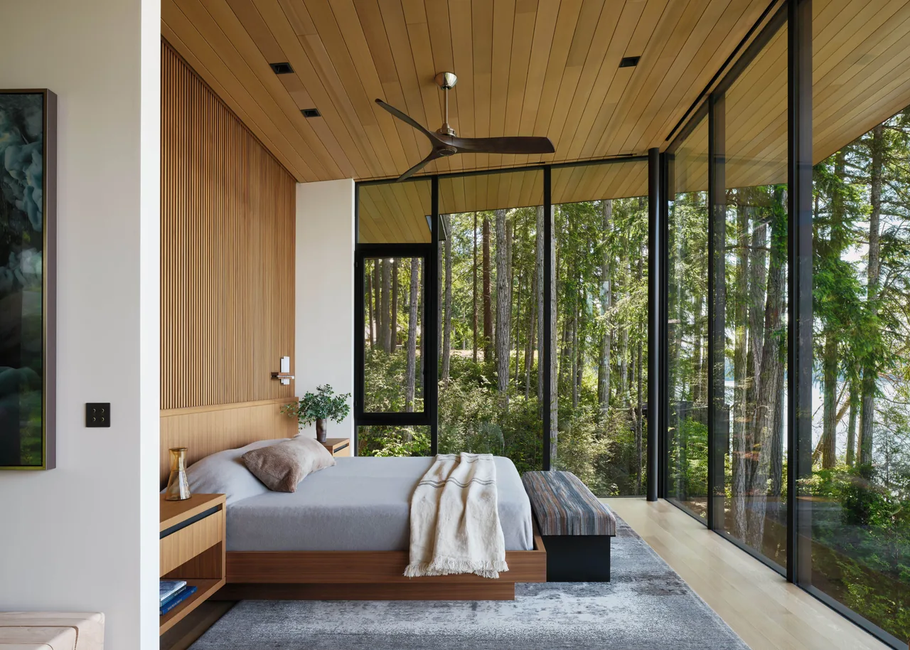 Bedroom with floor-to-ceiling window walls, wood-paneled ceiling, and a view of a wooded landscape including a lake beyond.