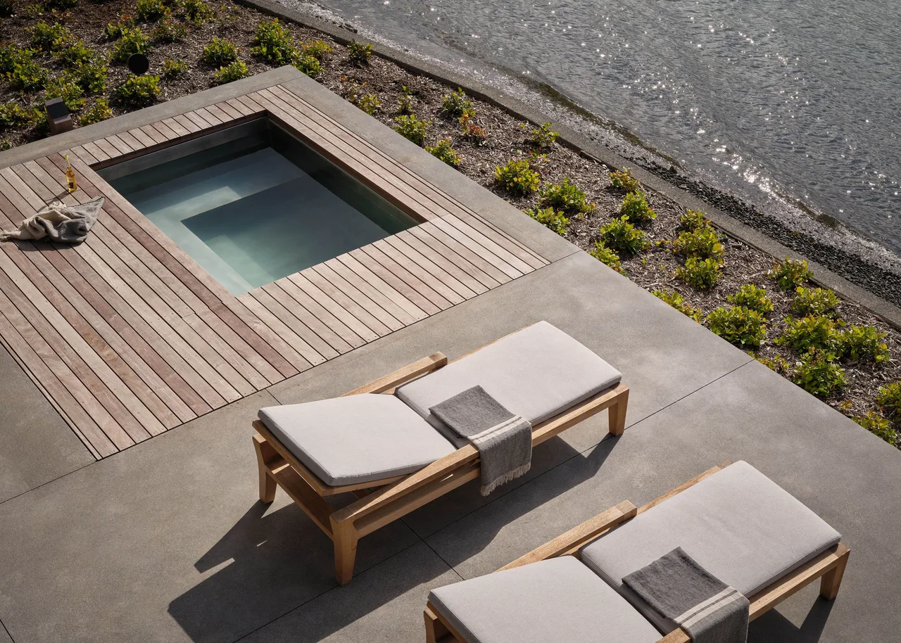 Lakeside concrete patio features a small sunken pool with adjacent wooden lounge chairs during sunset lighting.