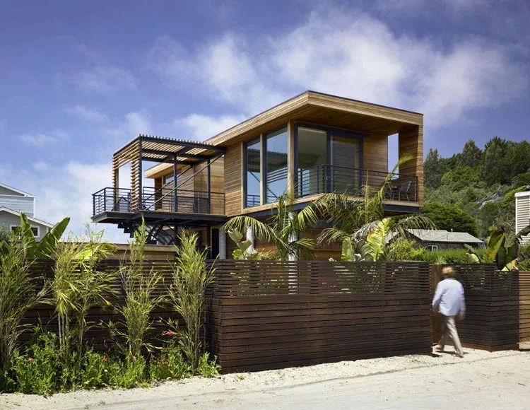 Modern two-story home with wood siding and large glass corner windows, featuring a balcony with black metal railing.