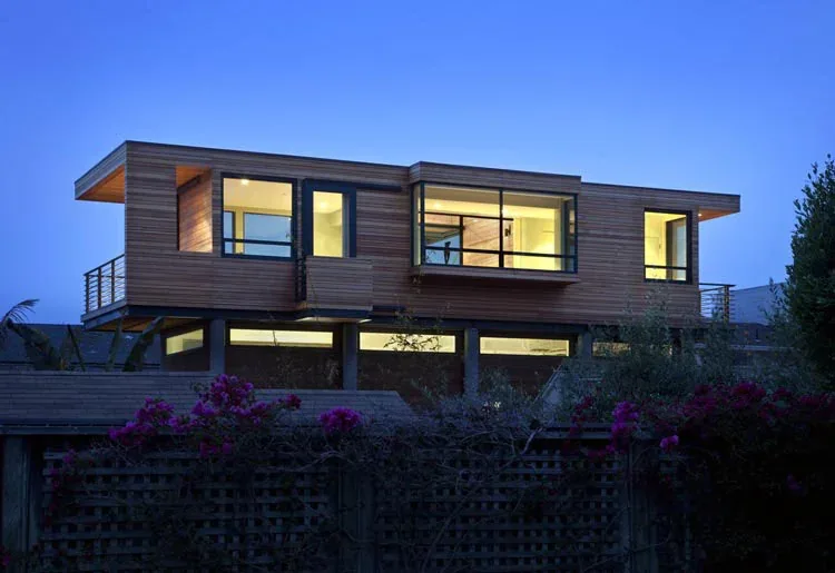 Modern elevated house with wood siding and large black window frames illuminated at dusk.